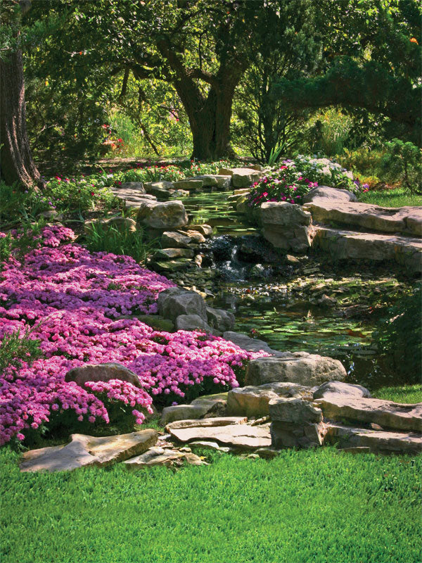 Lush garden with pink flowers, stone path, and trees surrounding a peaceful stream.