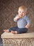 Baby sitting on a carved pedestal in front of an elegant vintage damask backdrop with gold and brown tones—suited for newborn photography, milestone portraits, and heirloom studio work.