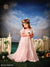 Young girl in pastel gown and floral crown posed in front of a fantasy-style Venice city backdrop with painted picket fence and daisies.