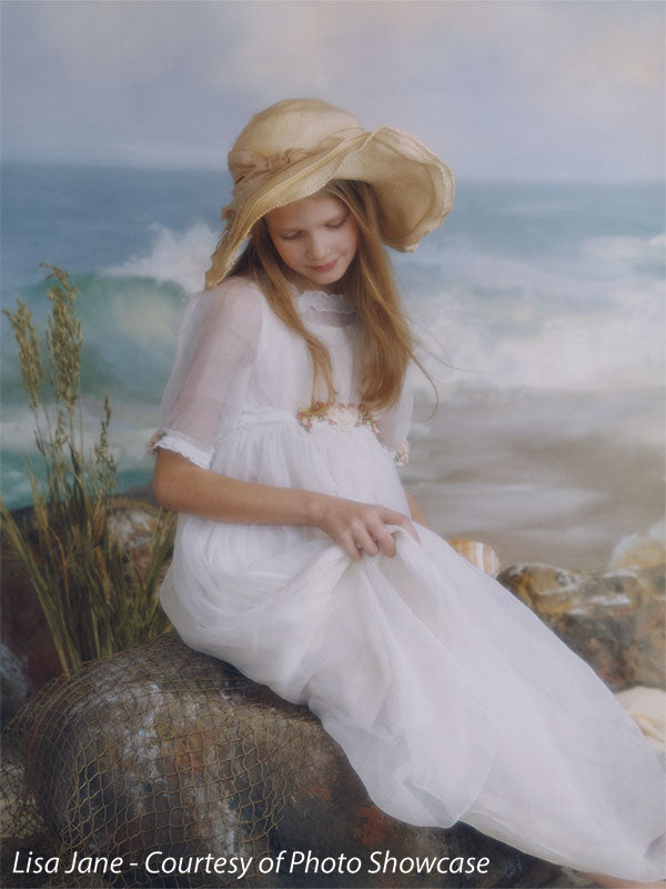 Young girl in vintage dress posed before a coastal beach backdrop, perfect for nostalgic children’s portraits and timeless editorial looks.
