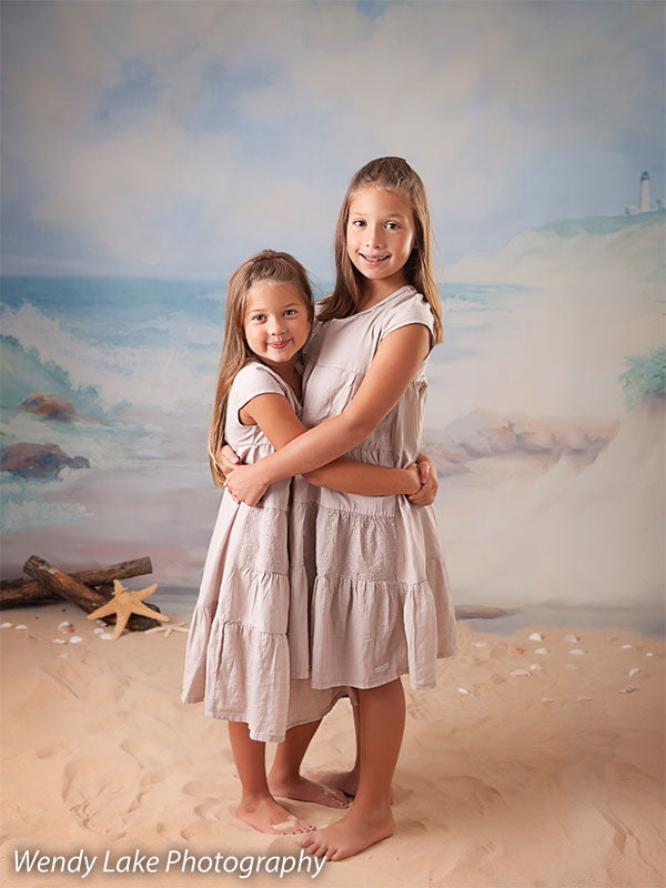 Sisters hugging barefoot on sand in front of a coastal lighthouse backdrop, ideal for natural family or children’s photography.