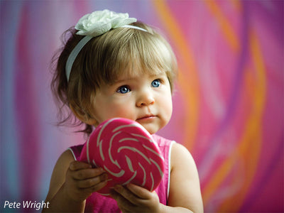 Toddler holding pink lollipop in front of vibrant abstract gift wrap-inspired backdrop, perfect for newborn or milestone studio sessions.
