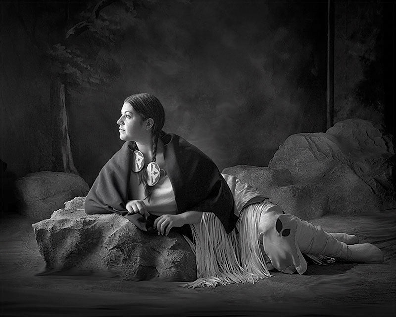 Black and white studio portrait featuring a model in native-inspired attire with a theatrical rock and fringe setup, captured against the swirling pink backdrop.