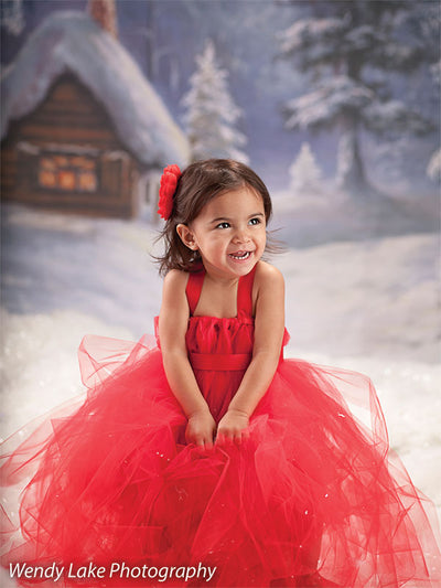 A printed snowy log cabin backdrop complements a child in a red holiday gown for a warm winter portrait setting.