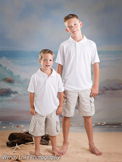 Two young boys in white posed on a sandy floor with waves and sky backdrop, suitable for school, family, or sibling portraits.