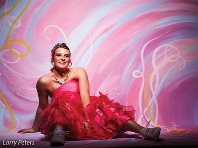 Young woman in bright pink dress seated against energetic pink swirl backdrop, great for styled senior or fashion portraits.