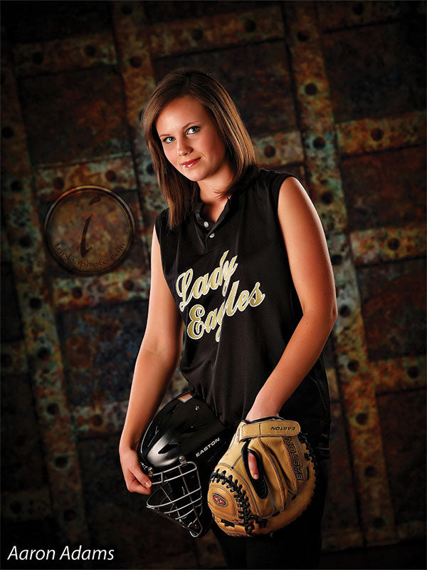 Female senior athlete posing in front of a weathered riveted backdrop with softball gear, enhancing bold textures for sports-themed portraits.