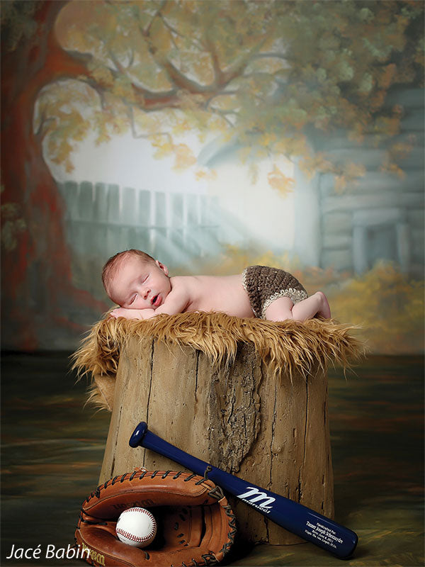 Printed backdrop showcasing a vintage log cabin and autumn tones, featured in a newborn photography setup with a baby model and baseball-themed props.