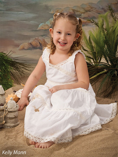 Girl seated in beach scene with sand and greenery in front of painterly shoreline backdrop, great for children’s and seasonal portraits.
