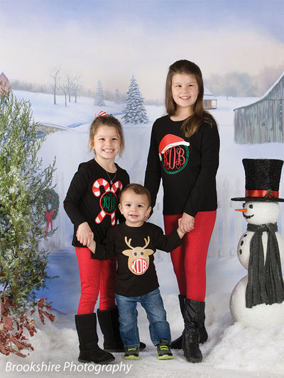 Three children in coordinated holiday clothing pose joyfully with a snowman in front of a snowy country backdrop.