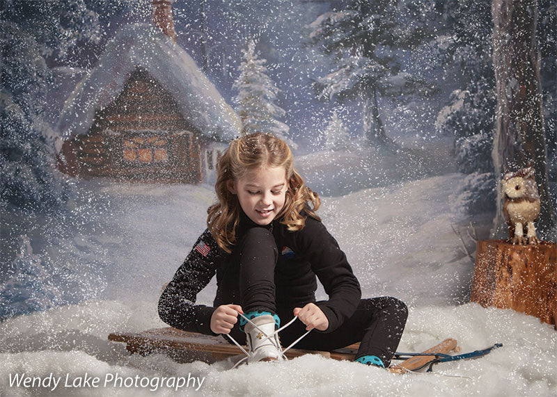 A snowy studio scene featuring a girl tying skates in front of a printed log cabin holiday backdrop.