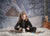 A snowy studio scene featuring a girl tying skates in front of a printed log cabin holiday backdrop.