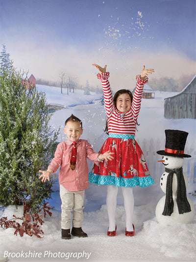 A cheerful holiday portrait with kids tossing snow in front of a printed snowy farm scene with barns and trees.
