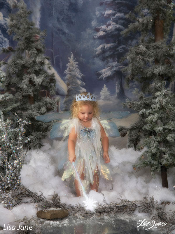 A young girl in fairy wings poses in a winter fantasy setup with the Holiday Cottage backdrop evoking a magical forest tale.